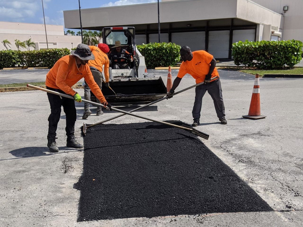 Three workers in orange shirts spread asphalt on a parking lot with rakes near traffic cones and machinery.