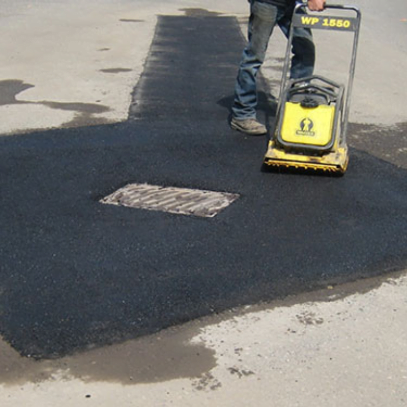 Person compacting freshly laid asphalt around a manhole cover on a road using a yellow machine.