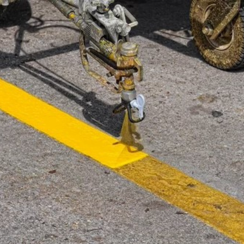 A machine paints a straight yellow line on a paved road, with wheels and spraying nozzle visible.