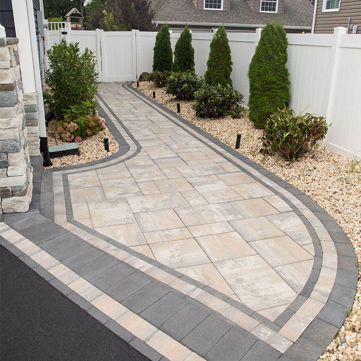 WLK-Alpine-Contemporary-South-Bay-Blend-3 Paved stone walkway bordered by plants, mulch, and a white fence in a landscaped yard.