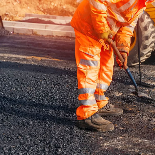 Worker in orange reflective gear spreading asphalt with a tool on a road construction site.