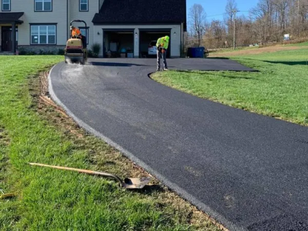 Two workers paving a new asphalt driveway in front of a house, with tools and equipment visible.