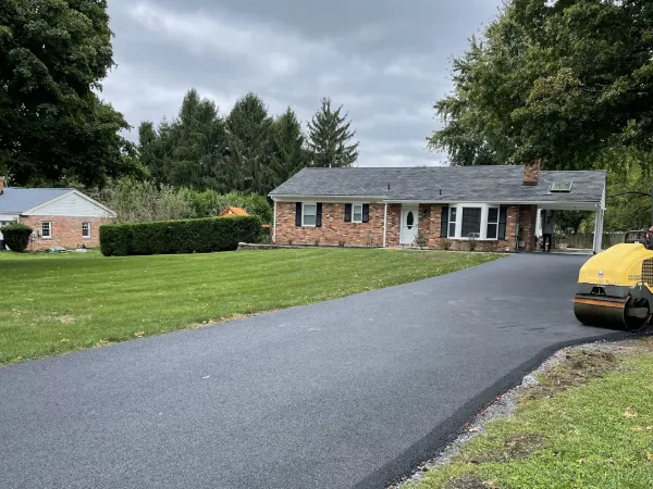 A freshly paved black asphalt driveway leads to a single-story brick house with a green lawn.