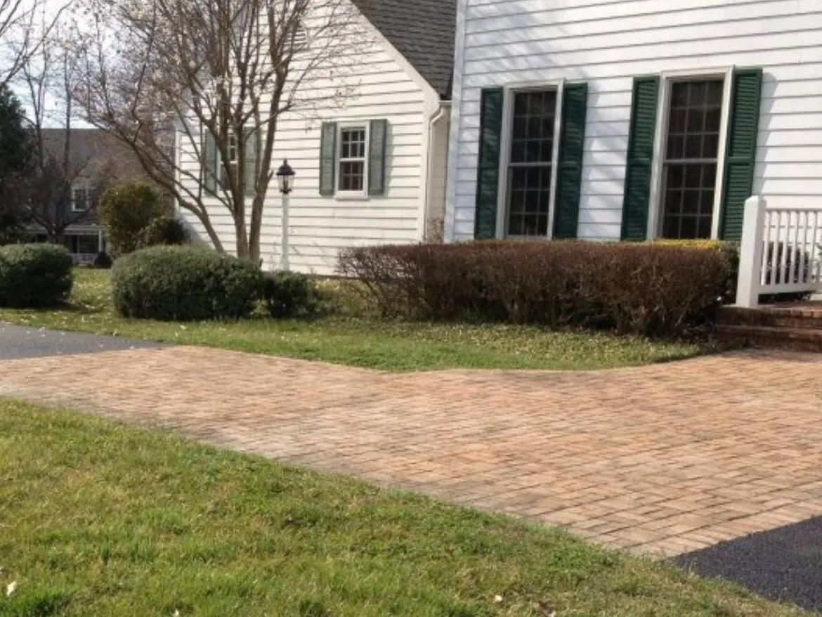 A brick driveway leads to a white house with green shutters and trimmed bushes in the yard.
