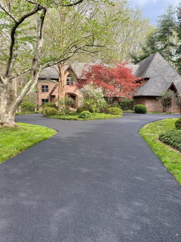 A brick house with peaked roofs, trees, and a curved driveway surrounded by green lawns and shrubs.