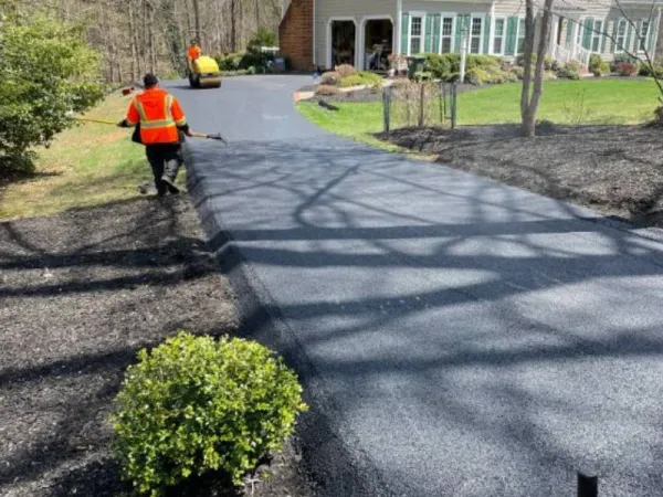 Workers in orange vests pave a black asphalt driveway in front of a house with a garden.