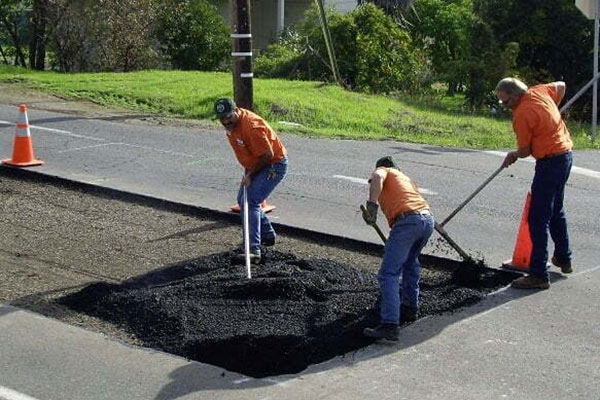 Three workers in orange shirts fill a large pothole with asphalt on a road near orange traffic cones.