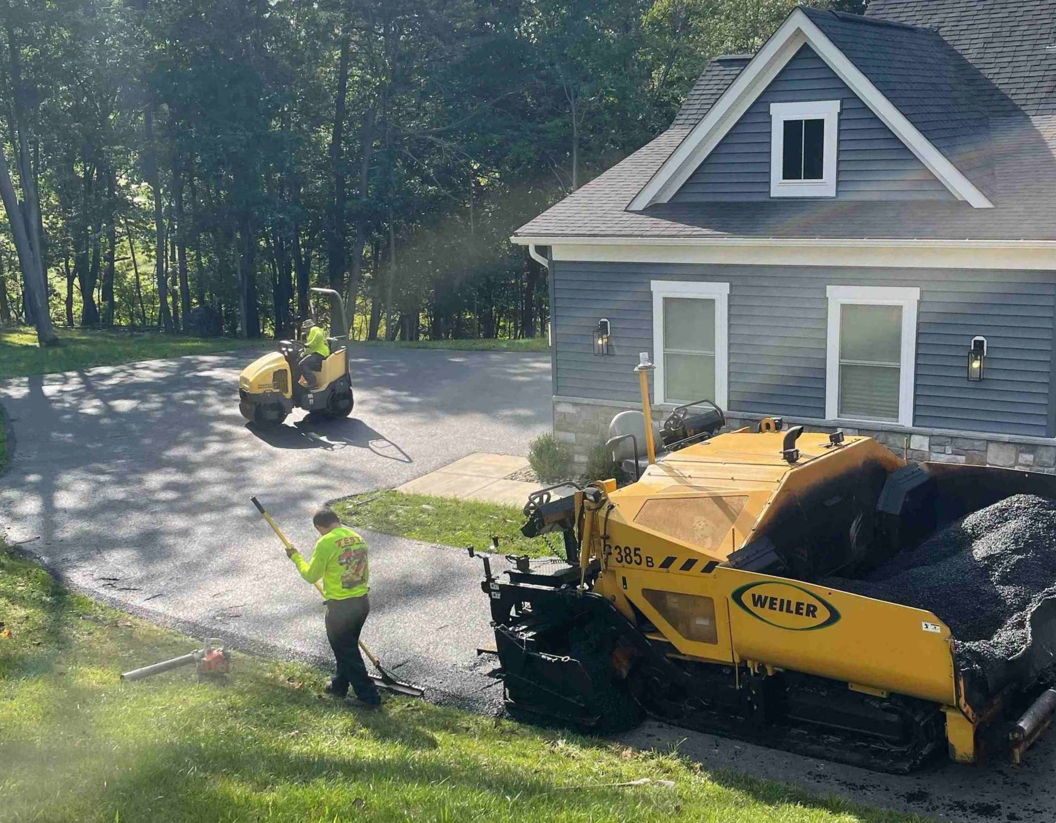 Two workers pave a driveway with heavy machinery beside a blue house surrounded by trees.