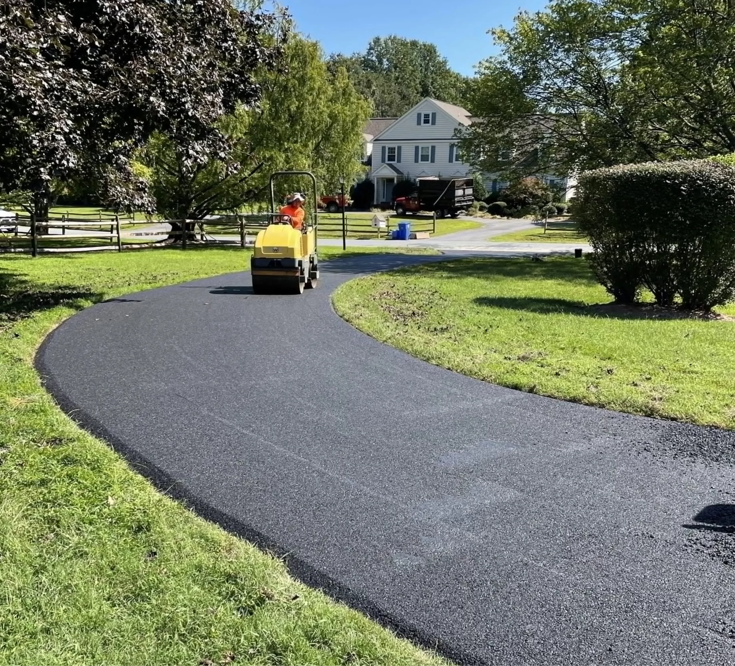 A worker uses a steamroller to pave a curved asphalt driveway in front of a house on a sunny day.