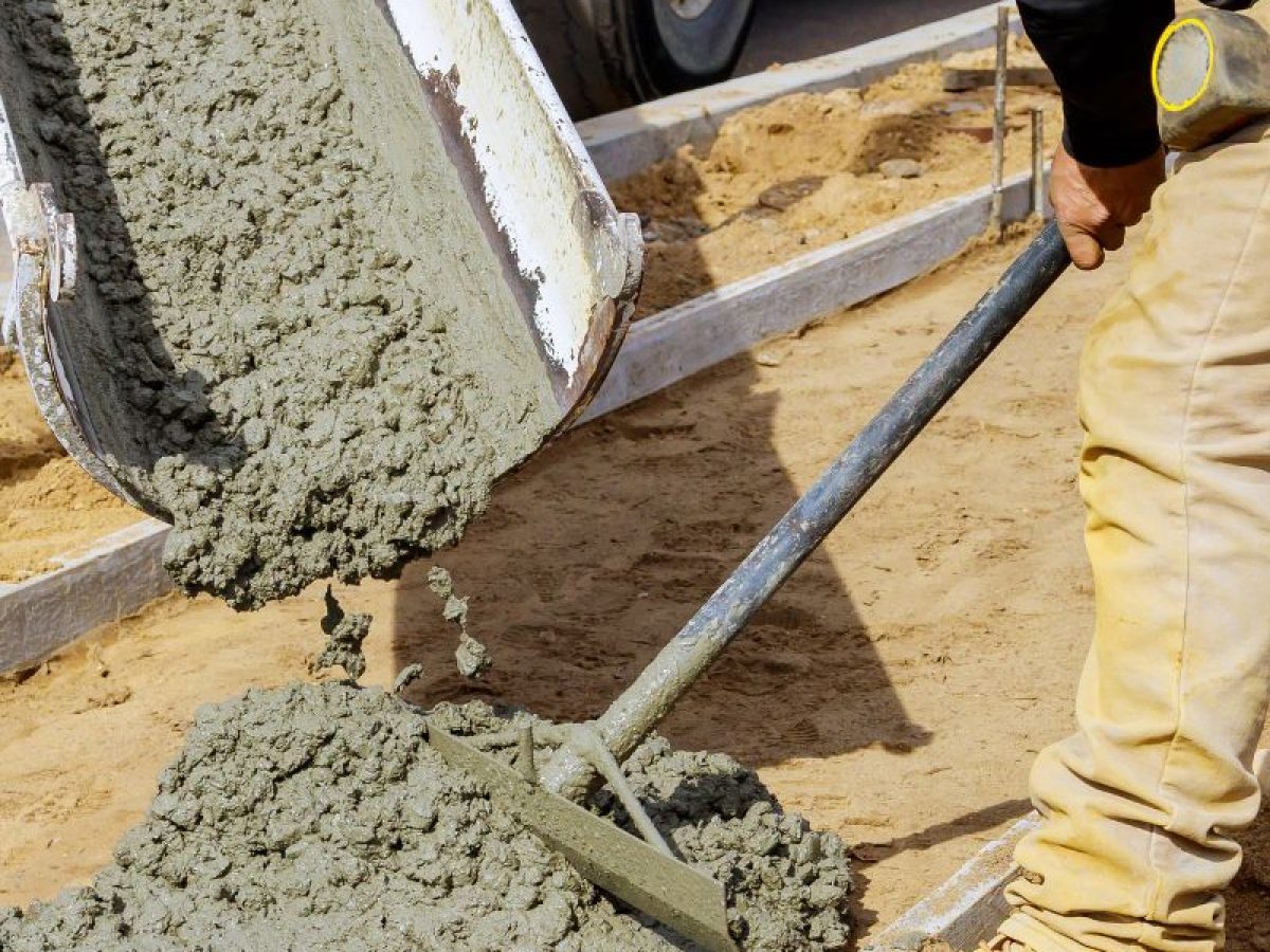 Wet concrete being poured from a chute onto sand, with a worker spreading it using a tool.