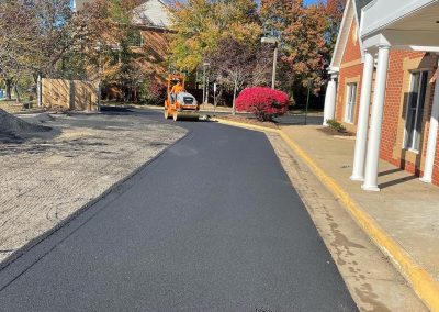 A worker drives a steamroller over freshly paved asphalt near buildings and autumn trees.