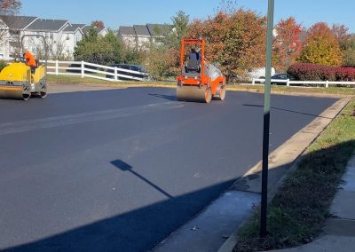 Two workers use orange steamrollers to pave a suburban street on a sunny day.