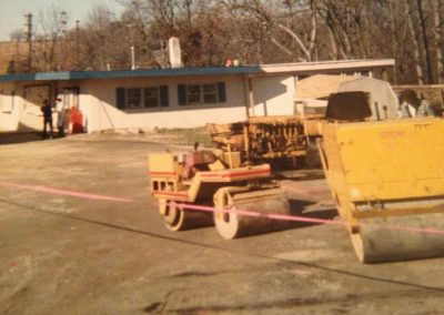 A small steamroller and construction equipment are parked near a single-story building, with trees in the background.