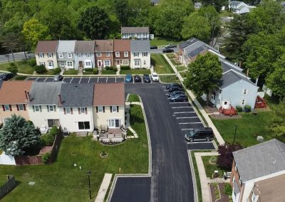 Aerial view of suburban houses, parked cars, and green trees on a sunny day.