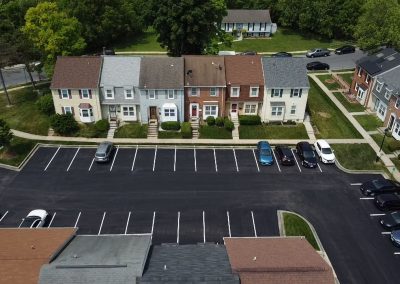 A row of townhouses faces a nearly empty parking lot, with a few cars parked, surrounded by trees and grass.