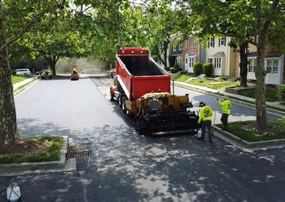 Workers repave a residential street with asphalt using heavy machinery on a tree-lined road.