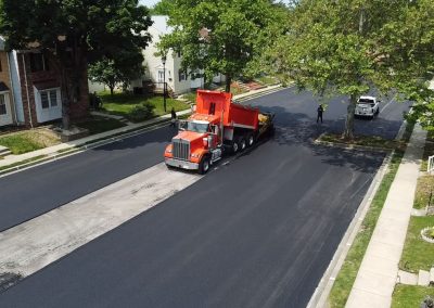 A red dump truck paves fresh asphalt on a residential street lined with houses and trees.