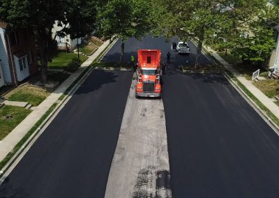 A red truck paves fresh asphalt on a suburban street lined with houses and trees.