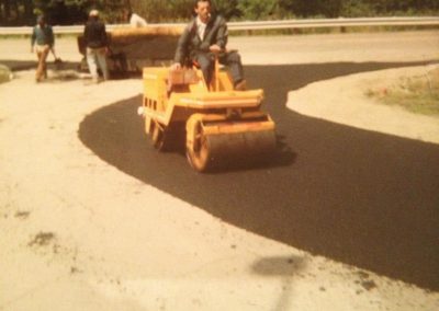 A man operates a steamroller on freshly paved asphalt, with two workers and equipment in the background.