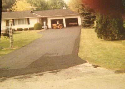 Two people work on paving a driveway in front of a single-story house with a garage and green lawn.