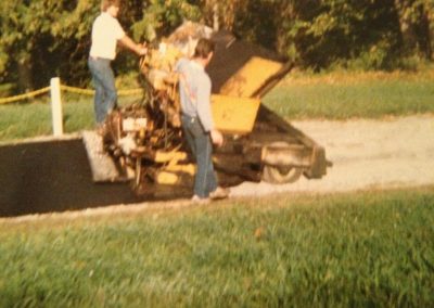 Two people operate a yellow asphalt paver on a road, surrounded by grass and trees.