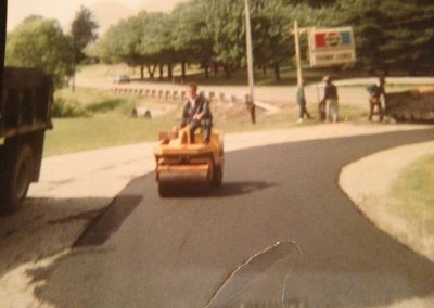 Man operates a small steamroller on newly paved road with workers and trees in the background.