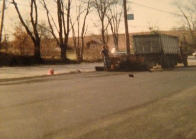 A truck parked by the roadside with a person standing nearby; leafless trees and a house in the background.