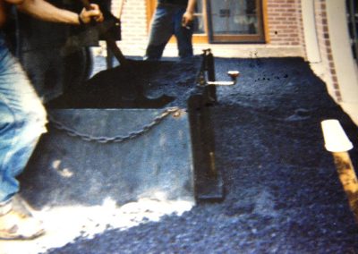 Two workers paving a surface with asphalt near a building, with paving equipment spreading black material.