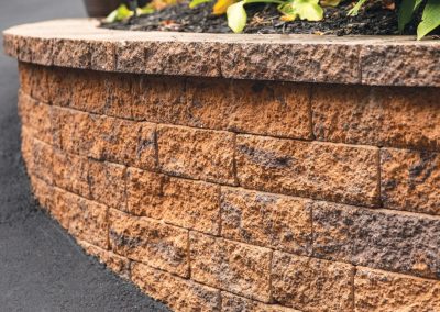 A curved, textured stone retaining wall borders a garden with green plants and mulch.