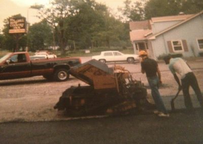 Two men operate a paving machine on a road, with a truck and a light-blue house visible in the background.