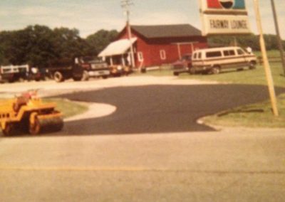 A red barn, vehicles, and a Farm Lodge sign with a Pepsi logo beside a newly paved road.