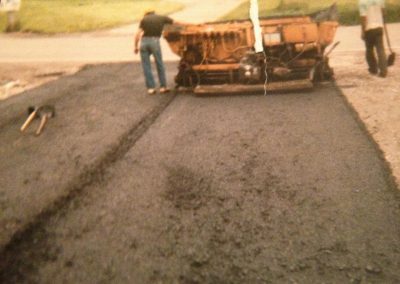 Two workers pave a road with asphalt using machinery; shovels lie on the freshly laid surface.