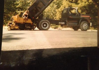 A dump truck unloads asphalt onto a road while a paving machine spreads it, trees in the background.