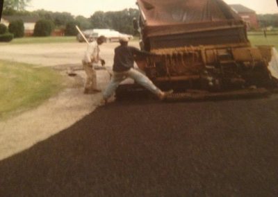 Two workers operate paving equipment while laying fresh asphalt on a road.