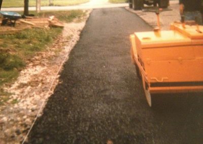 A yellow steamroller compacts fresh asphalt on a driveway under construction in a residential area.