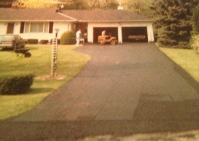 A house with a long driveway, a car, two people, and a yellow riding mower near an open garage.