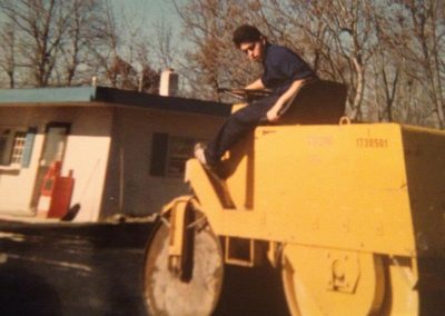 A person operating a yellow steamroller on a paved area near a small building with trees in the background.