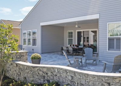 Backyard patio with gray chairs, potted plants, and a stone wall attached to a light gray house on a sunny day.