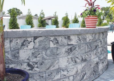 Curved stone wall with blue chairs and potted plants on top, outdoors on a sunny day.