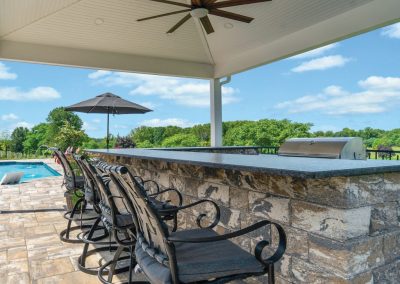 Outdoor patio bar with stone counter, chairs, ceiling fan, grill, and poolside view under a covered roof.