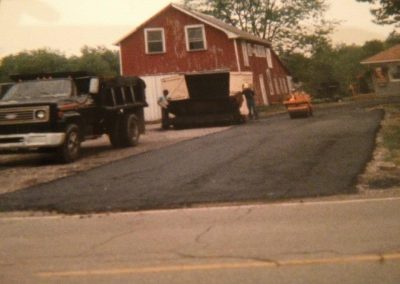 Workers pave a driveway with asphalt near a red barn and parked trucks on a rural property.
