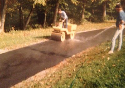 Person operating a yellow steamroller on a newly paved path, with another person standing nearby in a park.