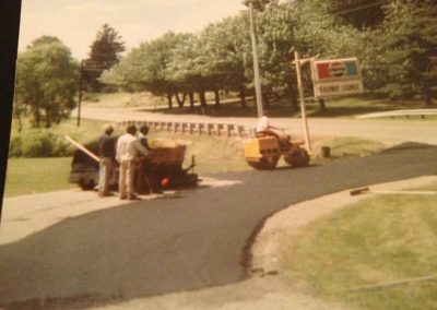 Four people paving a road; one operates a steamroller near a Pepsi Subnet Lounge sign, trees in background.