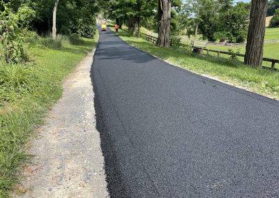 Fresh asphalt being laid on a rural road lined with green trees and grass under a blue sky.