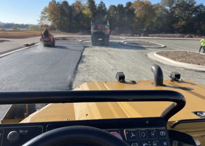 View from inside a construction vehicle, paving a road with other machines and workers visible ahead.