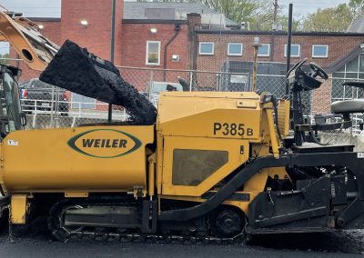 Yellow Weiler P385B asphalt paver spreading fresh asphalt near a brick building on a cloudy day.