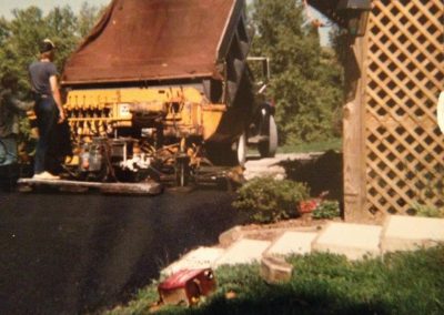 Two workers operate a yellow asphalt paving machine near a wooden lattice and a grassy yard.