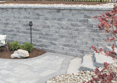 Gray stone patio with steps, curved retaining wall, landscaping rocks, and a leafy shrub in the foreground.