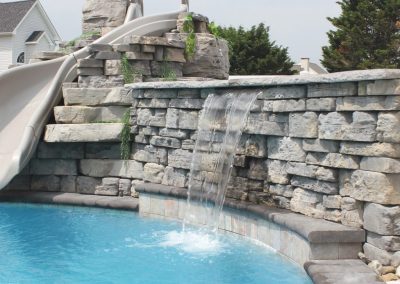Stone pool with a curved slide and a small waterfall flowing into the blue water, surrounded by greenery.
