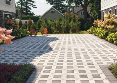 A spacious patio with geometric tile pattern, flanked by shrubs, and two wooden chairs in the distance.